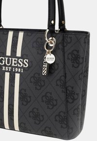 Borsa tote nera con strisce bianche e logo 'GUESS EST. 1981'. Presenta un portachiavi e charm decorativi attaccati al manico.