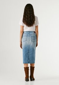 Vrouw met lang donker haar, gekleed in een wit topje met korte mouwen, een vervaagde blauwe denim midi-rok en bruine laarzen met hak, staande met de rug naar de kijker.