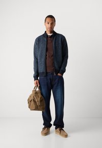 Jachetă bomber tricotată bleumarin cu un model herringbone, polo maro dedesubt, blugi închisi și o geantă bej cu mânere.