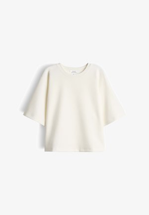 Crèmekleurig oversized T-shirt met korte mouwen en een ronde hals, met een klein label aan de binnenkant van de hals, tegen een witte achtergrond.