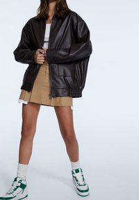 Brązowa bomberka z sztucznej skóry o oversize'owym kroju, z zamkiem z przodu, bocznymi kieszeniami, noszona na beżowej plisowanej spódnicy i białych-zielonych sneakersach.