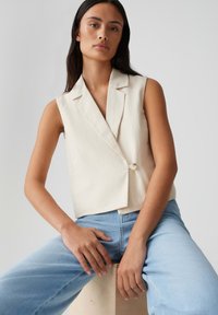 Blazer senza maniche beige con chiusura a un bottone, colletto scollato e texture liscia, abbinato a jeans azzurri chiari.