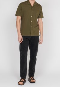 Olivgrön kortärmad skjorta med krage och knappstängning, tillsammans med svarta jeans och bruna sandaler. Avslappnad passform och slät textur.