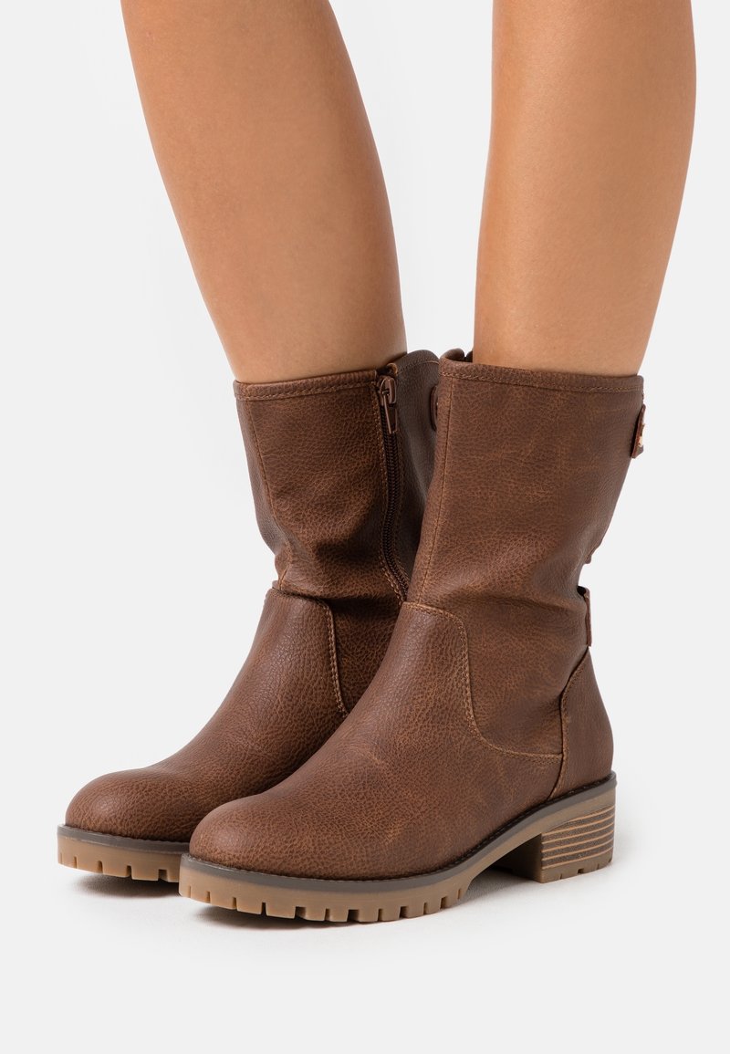 Bottes en cuir marron mi-mollet avec fermetures éclair latérales, finition texturée et talons bas empilés portées sur des jambes nues sur fond blanc.