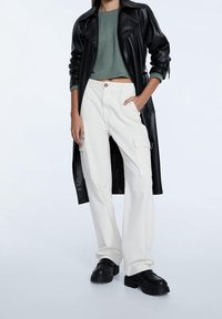 Manteau long en cuir synthétique noir, pull court côtelé vert, pantalons cargo crème avec poches, chaussures à plateforme noires. Design minimaliste et fonctionnel.