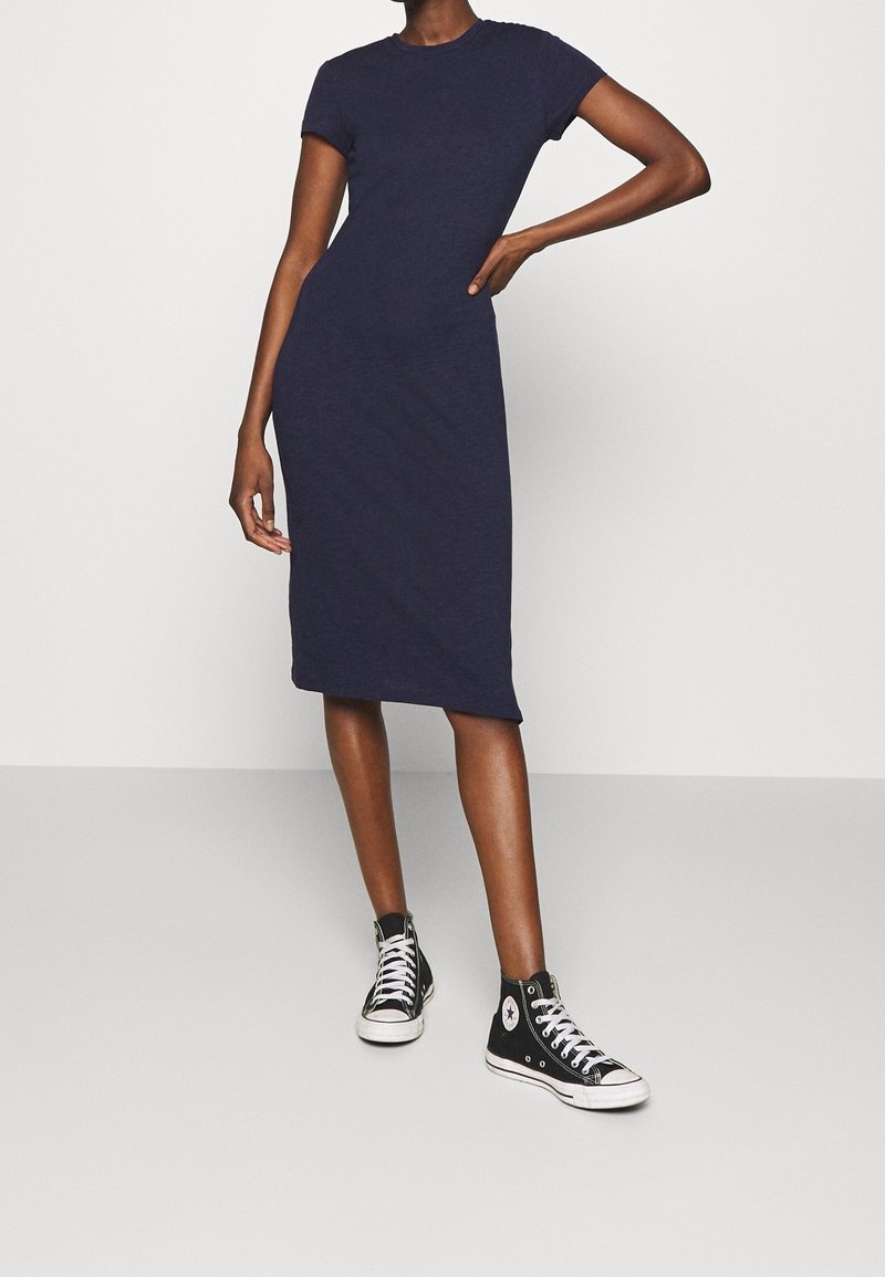Femme portant une robe bleu marine cintrée jusqu'aux genoux et des baskets montantes noires, debout avec une main sur la hanche.