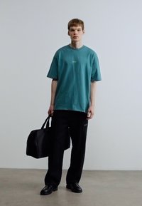 Πράσινο-μπλε υπερμεγέθες μπλουζάκι με πράσινο λογότυπο, σε συνδυασμό με φαρδιά μαύρα παντελόνια και μαύρα δερμάτινα παπούτσια, κρατώντας μια μαύρη ταξιδιωτική τσάντα.