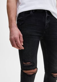 Primer plano de una persona que lleva vaqueros negros desgastados y una camisa blanca, con una mano relajada a su lado.