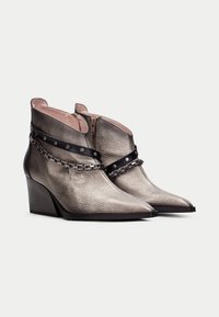 Botines metálicos plateados hasta el tobillo con punta afilada y tacón de cuña. Presentan una correa negra con tachuelas y detalle de cadena. Acabado en cuero texturizado.