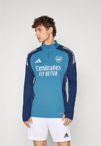 Teal and navy long-sleeve sports top with zip collar, featuring white "Emirates Fly Better" logo and three white stripes on shoulders.