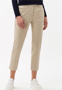 Beige cropped broeken in een soepele stof, met zakken aan de voorkant en een knoopsluiting, gecombineerd met witte sneakers voor een casual look.