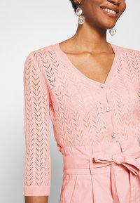 Femme portant un cardigan en maille rose à motif chevron et un pantalon taille haute assorti ceinturé de tissu, avec des boucles d'oreilles pendantes transparentes.