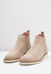 Pár béžových semišových kotníkových bot s elastickými bočními panely a bílými podrážkami, vystavených na bílé ploše na jednobarevném pozadí.