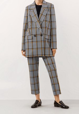 Blazer oversize à carreaux noir, blanc et moutarde avec fermeture à double boutonnage ; pantalon assorti sur mesure et mocassins noirs brillants.