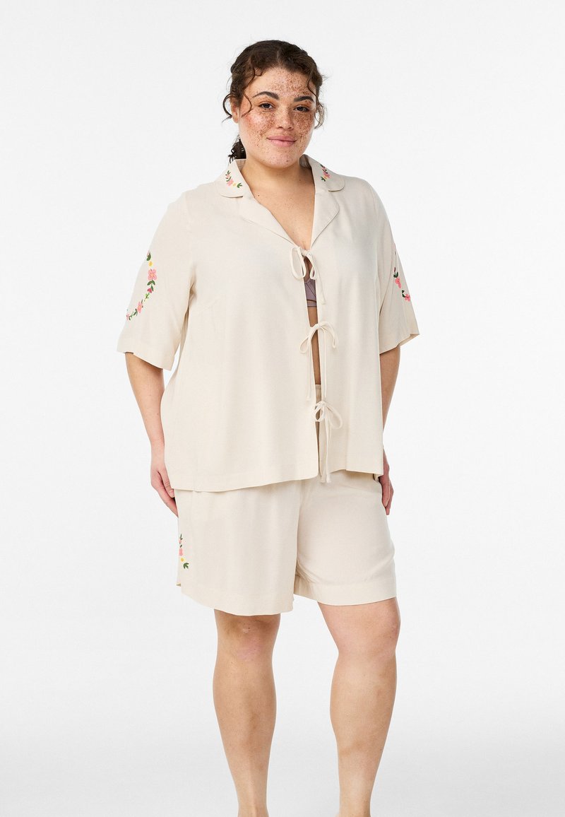 Béžový súprava na voľný čas zahŕňa krátky rukáv, top so zaväzovaním vpredu s kvetinovou výšivkou na rukávoch a zodpovedajúce šortky s podobnými detailmi.