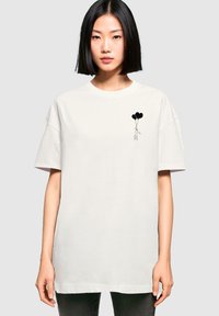 Witte oversized t-shirt van katoen, met een zwarte lijntekening van een figuur die drie zwarte ballonnen vasthoudt op de linkerkant van de borst.