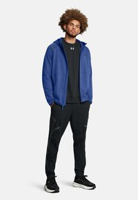 Blaue Zip-Hoodie mit grauen Akzenten, kombiniert mit einem schwarzen T-Shirt und schwarzen Hosen mit Seitentaschen. Schwarze Sneakers runden das Outfit ab.