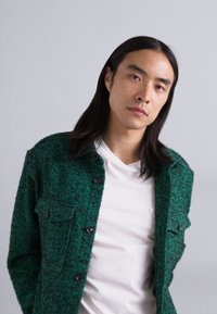 Veste texturée verte avec des fermetures à bouton et une poche poitrine, superposée à un T-shirt blanc, présentant une coupe moderne et un tissu doux.