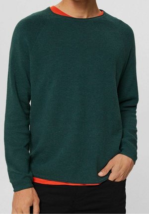 Uomo che indossa un maglione a maniche lunghe verde scuro sopra una camicia arancione, con una mano in tasca, mostrato dal collo ai fianchi.
