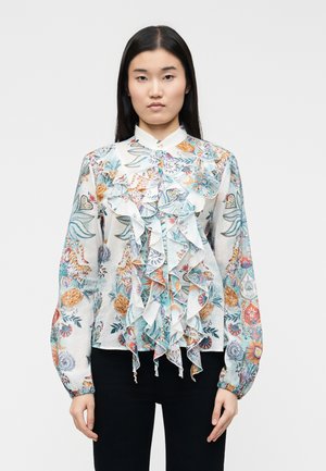 Azjatka o długich czarnych włosach, ubrana w białą bluzkę z kolorowym kwiatowym i zwierzęcym wzorem oraz spływającymi falbanami z przodu, w połączeniu z czarnymi spodniami.