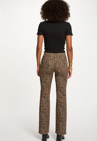 Vrouw met krullend haar, gekleed in een zwart t-shirt met korte mouwen en bruin leeuwenkleurig rechte broek met zwarte haklaarzen, staande met de rug naar de camera.