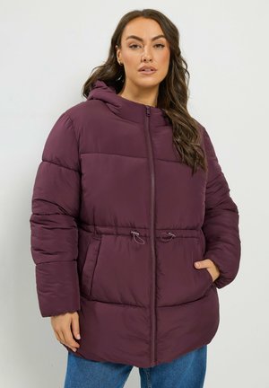 Femme portant une veste matelassée violette avec capuche, fermeture éclair et cordon à la taille, debout devant un fond clair uni.