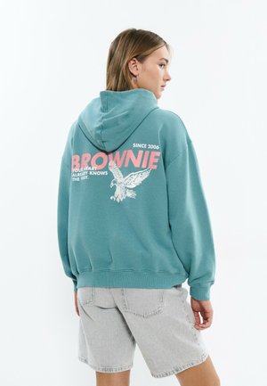 Jeune femme portant un sweat à capuche bleu sarcelle avec le texte rose "BROWNE" et un motif d'aigle, associé à un short en jean gris clair, vue de profil.