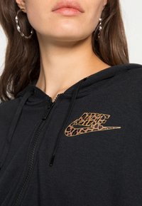 Zwarte hoodie met ritssluiting en een Nike-logo in luipaardprint. Voorzien van trekkoorden en een zachte textuur, met een close-up van de halslijn.