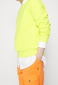 Neon žlutá mikina se žebrovanými manžetami, vrstvená přes bílou košili s knoflíky. V páru s oranžovými kalhotami s kapsami a emblémem.