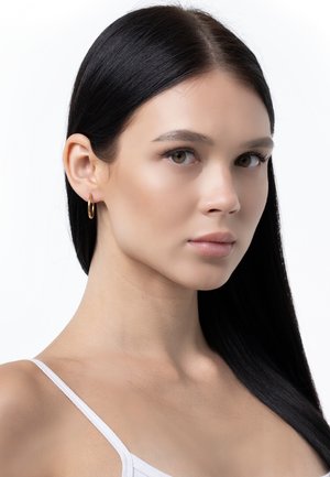 Giovane donna con lunghi capelli neri che indossa una maglietta bianca e un orecchino a cerchio dorato, che guarda leggermente di lato su uno sfondo bianco.