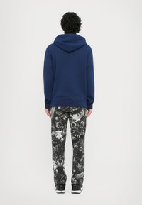 Felpa blu con cappuccio e lacci, abbinata a pantaloni della tuta neri e bianchi con motivo. L'outfit presenta una vestibilità comoda e un design casual.
