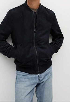 Personne portant une veste bomber noire zippée, les mains dans les poches et un jean bleu clair, debout devant un fond blanc uni.