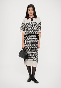 Conjunto de malha em bege com padrões florais pretos, mangas curtas e um colarinho borgonha. Combinado com uma mala preta e collants pretos.