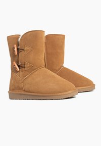 Tan suède enkelboots met een ronde neus, voorzien van twee driehoekige accenten en houten knopen aan de zijkant. Zachte binnenvoering. Rubberen zool.