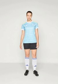 Kvinne som står iført lys blå fotballdrakt, svarte shorts, hvite knelange sokker med blå striper og svarte fotballsko mot en ensfarget bakgrunn.