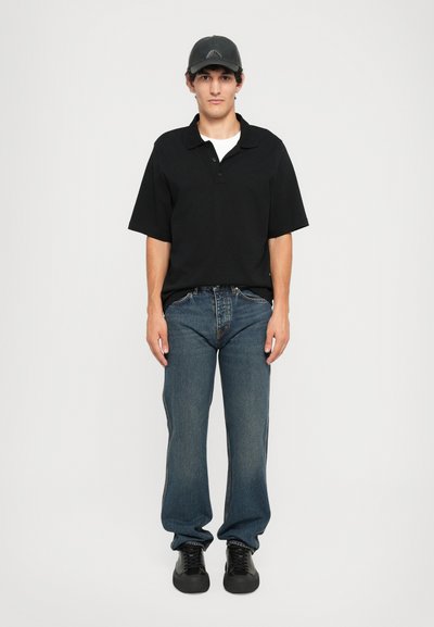 Polo shirt noir à manches courtes, jean droit bleu, baskets noires et casquette noire portés par un mannequin masculin debout sur un fond uni.