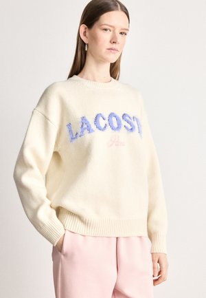 Kermaviininvärinen neule, jossa on ribbireunat hihoissa ja helmassa, ja jossa on pörröiset siniset "LACOSTE"- ja vaaleanpunaiset "Paris"-logot. Sileä pinta, rento istuvuus.