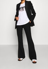 Svart blazer med satinlapeller, vit T-shirt med grafiskt tryck, svarta utsvängda byxor och vita sandaler med remmar.