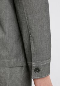 Gray long-sleeve shirt fabric with a textured finish, featuring a cuff detail with a black button and contrasting white stitching.