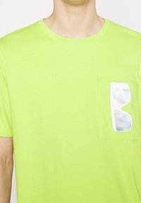 Une personne portant une chemise à manches courtes vert vif avec une petite poche sur la poitrine, tenant des lunettes de soleil iridescentes.