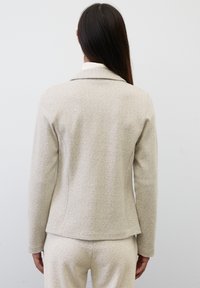 Béžový herringbonový blazer s přiléhavým střihem, zářezovým límečkem a jemnou texturou, spárovaný s odpovídajícími béžovými kalhotami.