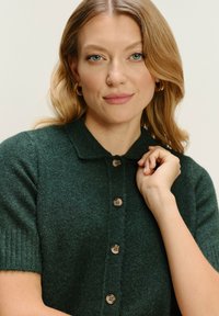 Dunkelgrüne Strickjacke mit Kragen, kurzen Ärmeln und vierknöpfigem Frontverschluss. Weiche Textur mit dezenten Rippungen an den Ärmeln.