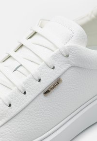 Zapatilla de cuero blanca con superficie texturizada, puntera redonda y cordones blancos planos. Presenta una pequeña placa de logo metálico en el lateral.