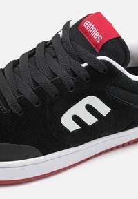 Black suede skate shoes with white logo, featuring black laces, red accents, a textured upper, and a white rubber sole with a red tread.