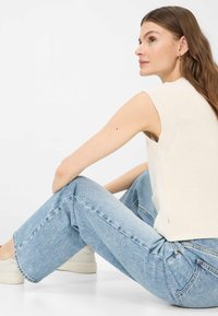 Donna con capelli castano lunghi che indossa un top lavorato a maglia senza maniche color crema, jeans azzurri chiari, scarpe da ginnastica bianche, seduta con le ginocchia piegate su un pavimento bianco.