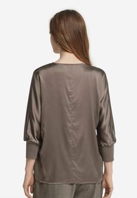 Blusa in satin bronze con maniche a tre quarti, scollo a girocollo e leggere arricciature sulle spalle. Tessuto liscio e vestibilità rilassata.
