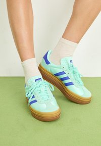 Mintgröna sneakers med blå accenter, med en texturerad häl, vita snören, en tjock brun sula och tre blå ränder på sidorna.