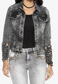 Musta ja harmaa farkkutakki, jossa on kulutettuja, pitsisiä yksityiskohtia hihoissa ja taskuissa. Yhdistetty vaaleanharmaisiin farkkuihin, joissa on strassikoristeita.