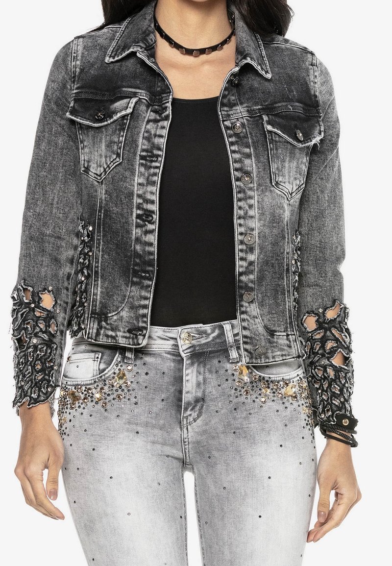 Musta ja harmaa farkkutakki, jossa on kulutettuja, pitsisiä yksityiskohtia hihoissa ja taskuissa. Yhdistetty vaaleanharmaisiin farkkuihin, joissa on strassikoristeita.