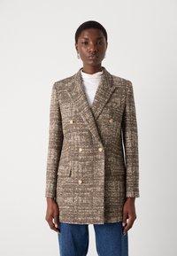 Doppelreihige Blazer aus braunem Fischgrätenmuster, mit zwei Fronttaschen und hellen Knöpfen. Kombiniert mit einem weißen Rollkragenpullover.